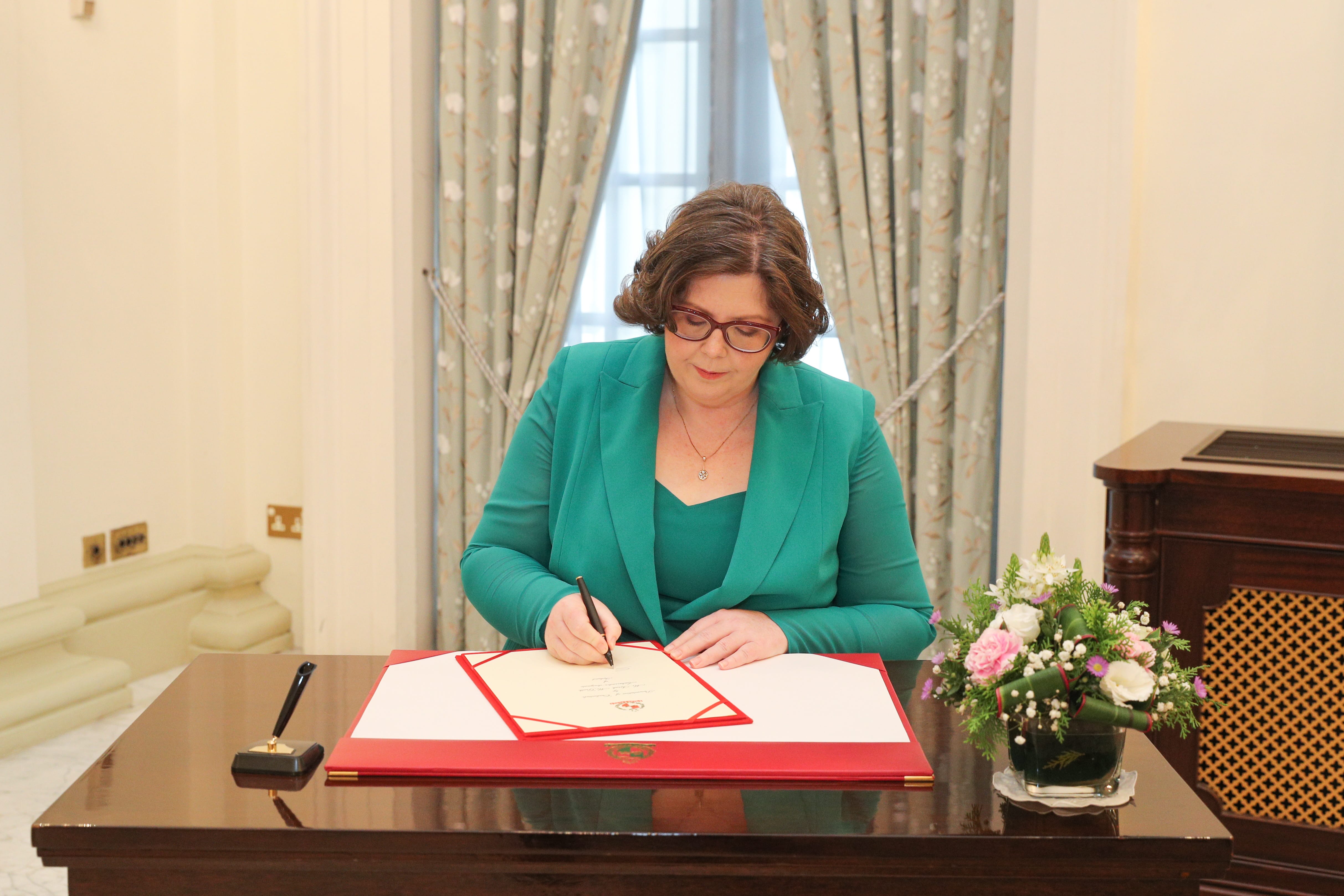 Woman in green suit signing document at desk with pen. Flowers to the side.